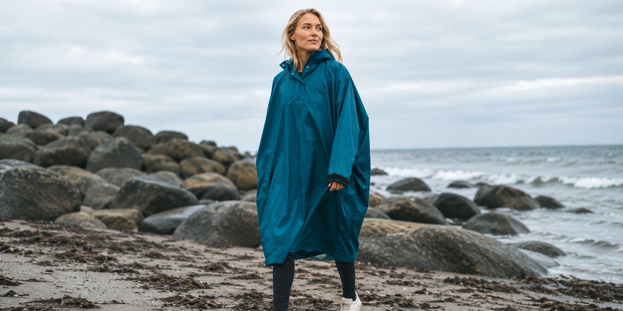 Elkline Unisex Poncho in der Farbe Petrol. Modell geht am Strand spazieren. Bereit für den nächsten Schauer